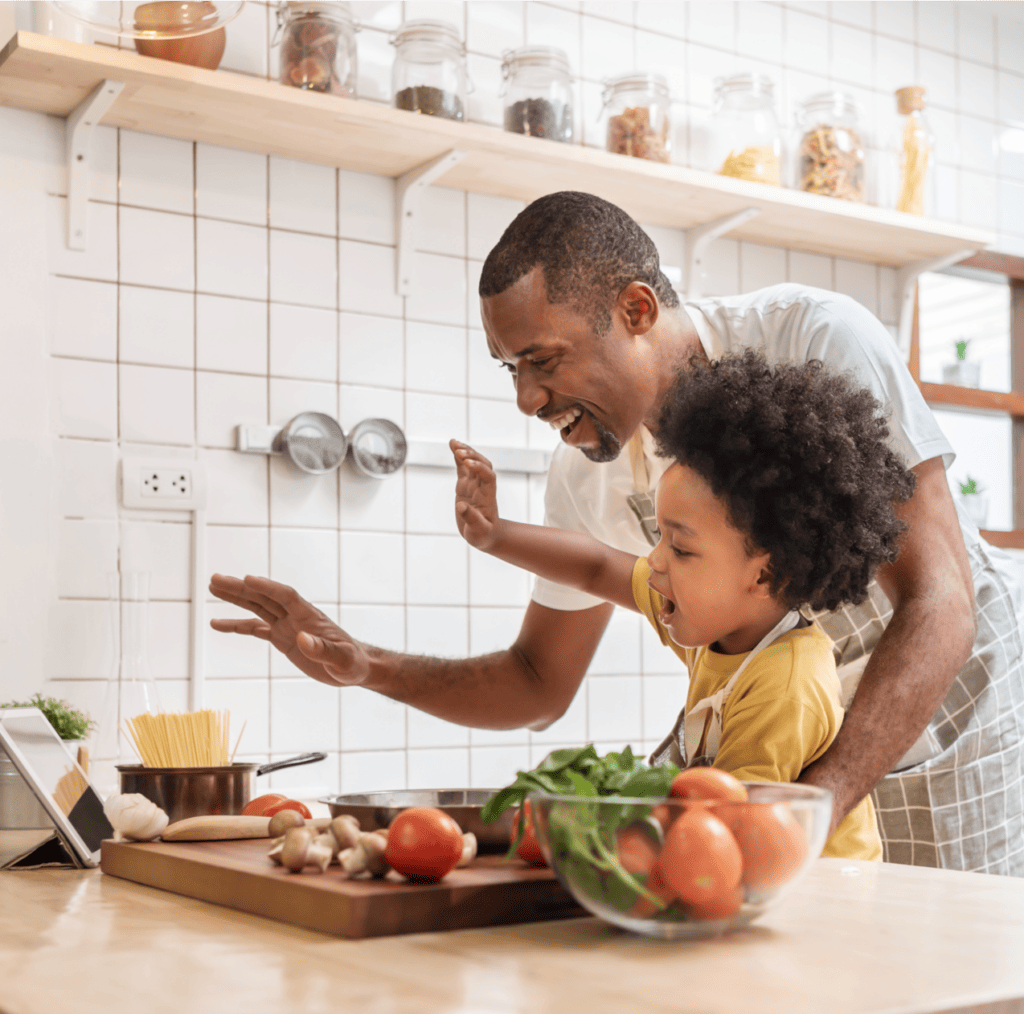 Black father and young son making video call with a digital tablet and waving hands laughing talking with family while cooking in the kitchen.