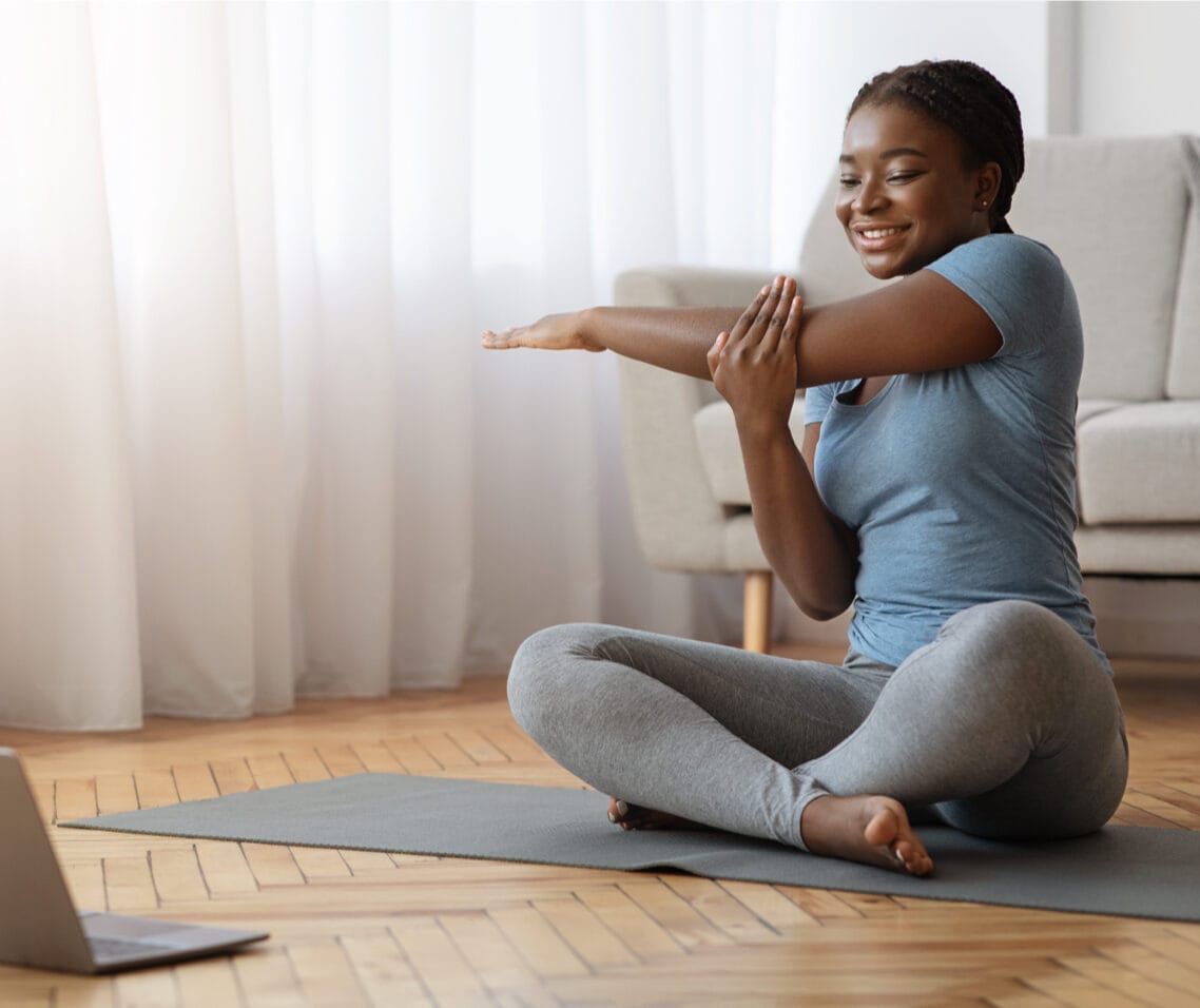 Cheerful black woman stretching in front of laptop, doing home workout in living room.