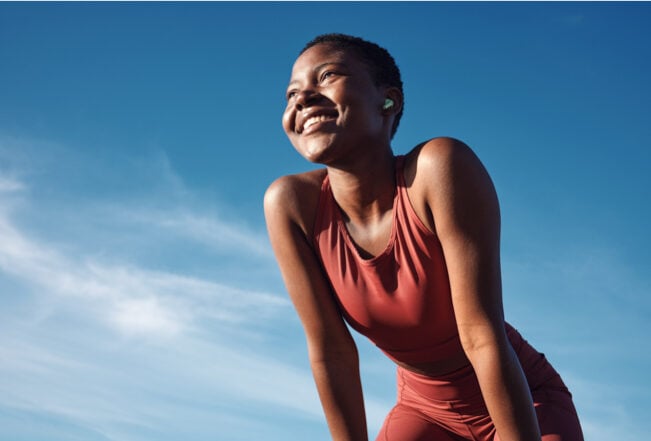 Low angle of black woman smiling after running, blue sky behind her.