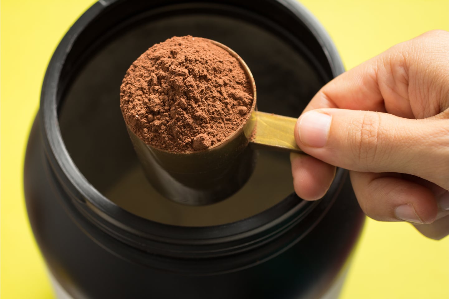 Close up of hand scooping chocolate protein powder out of container with yellow background.