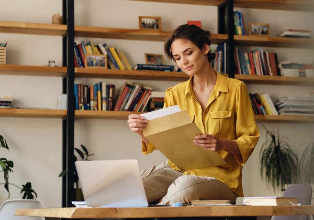 Young smiling woman in a yellow shirt sitting on a desk with papers and laptop working in a modern office.