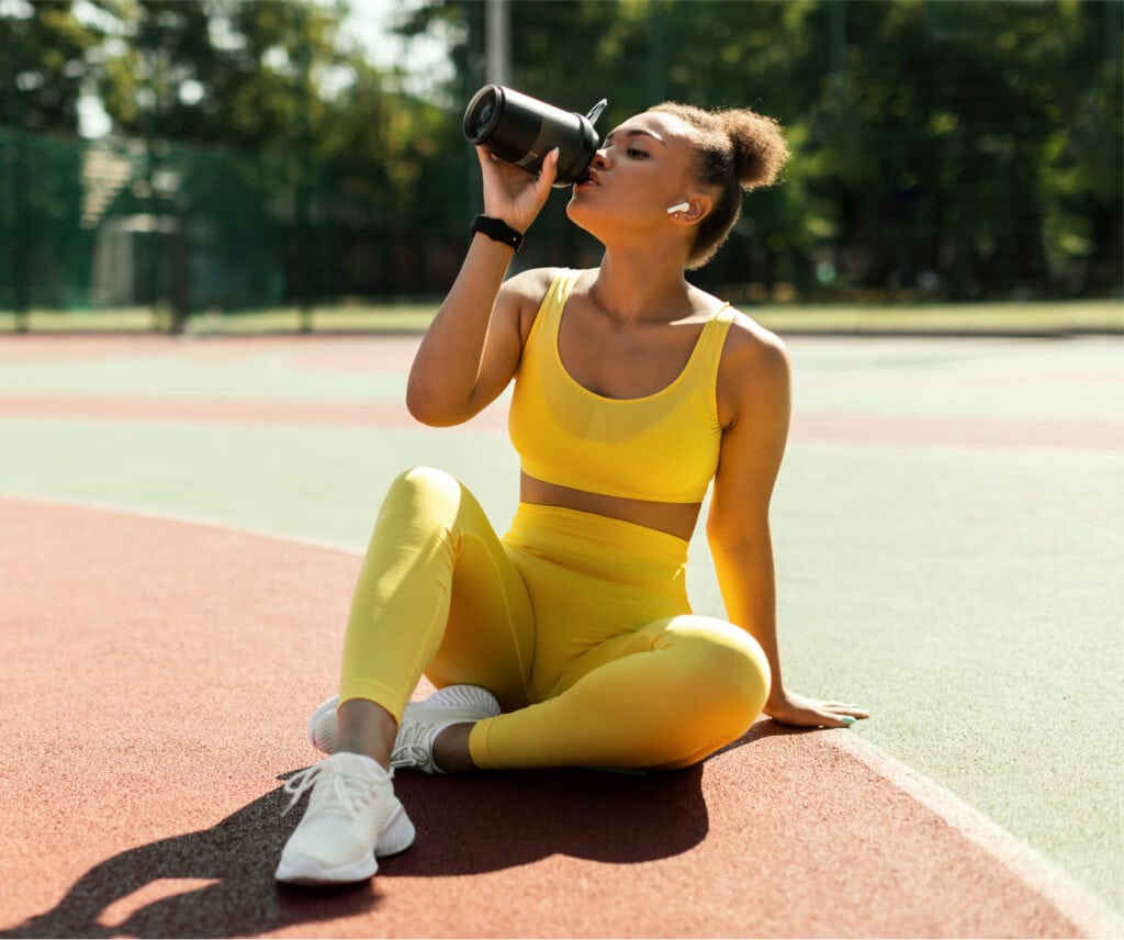 Fit black woman in wireless earbuds sitting on basketball court ground, drinking from shaker, taking break after outdoors exercise.